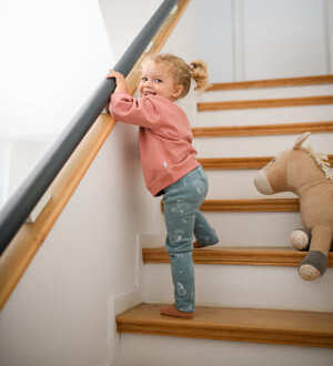 Ein kleines Mädchen in einem rosa Pullover und Jeanshose klettert lächelnd eine Holztreppe hinauf.